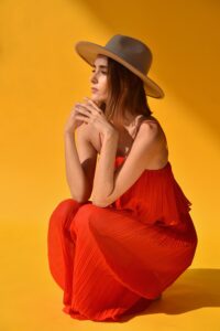 A woman in a red dress sitting cross-legged against a yellow background, looking contemplative, with a grey hat on her head.